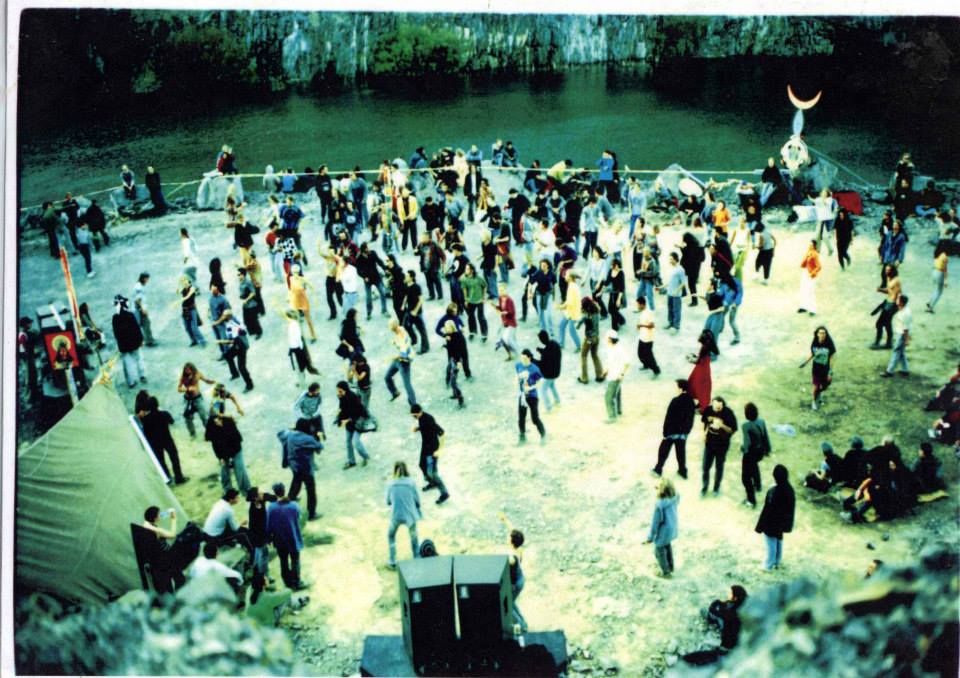 Dancers spread across a quarry floor beside still water, with a crescent-moon sculpture at the far edge