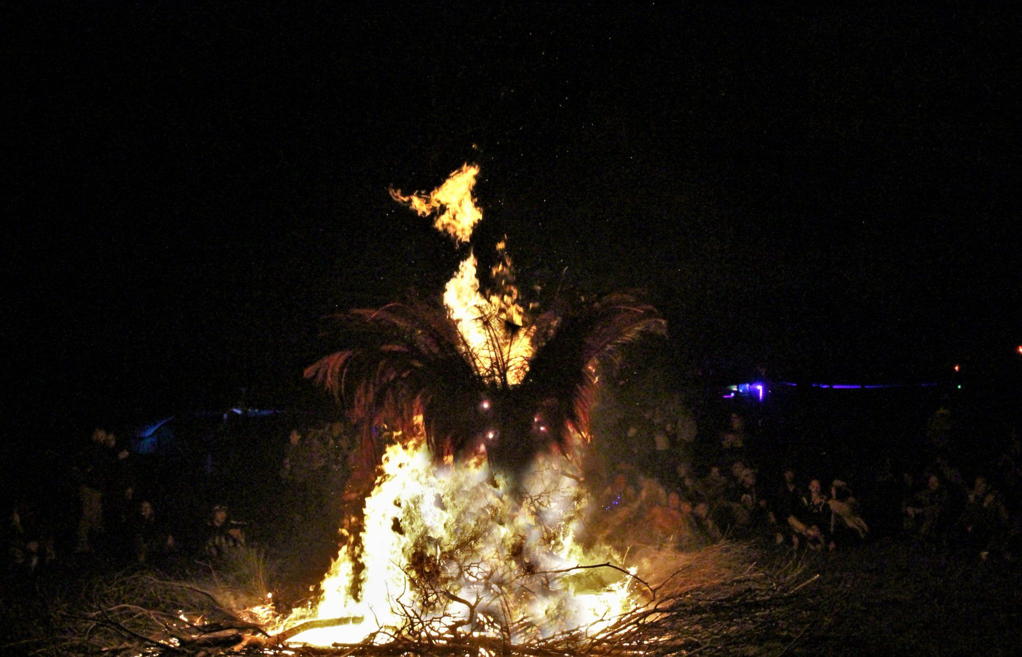 A burning structure at the centre of a wide circle of people standing in the dark