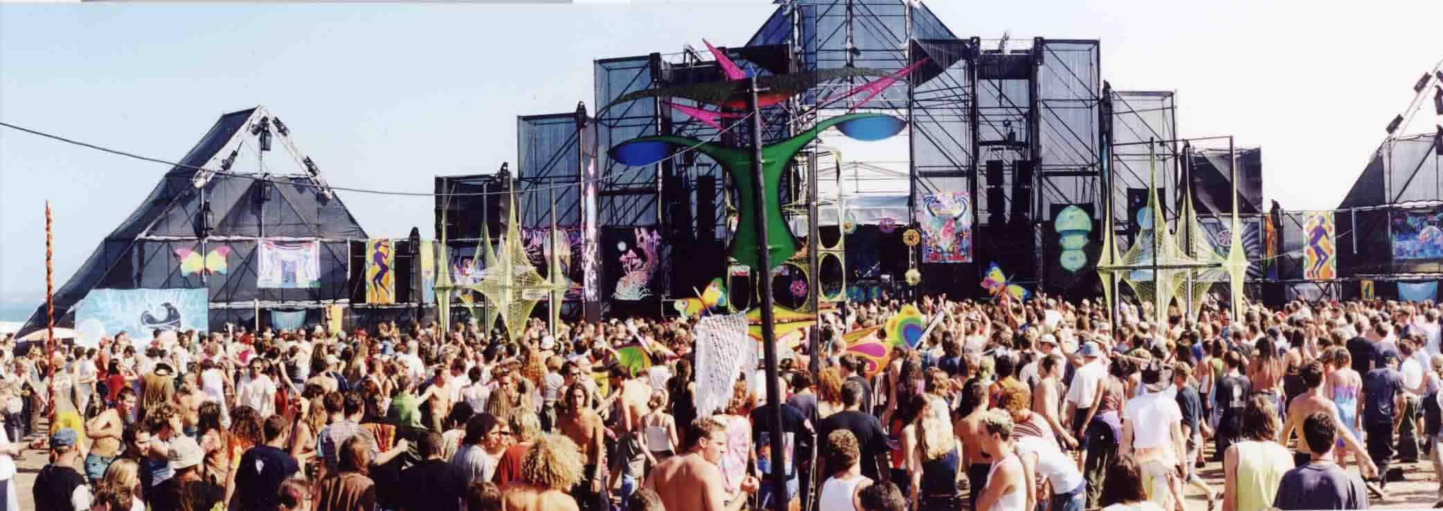 A large daytime crowd in front of a scaffolded beach stage hung with painted banners and net sculptures