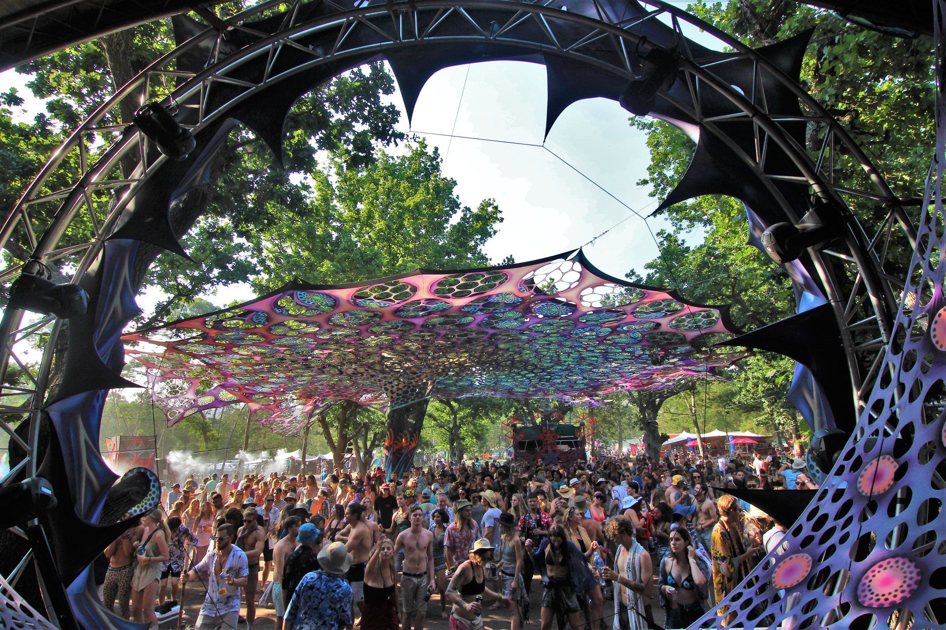 A daylight crowd dancing beneath a patterned fabric canopy strung between trees at a Vortex festival