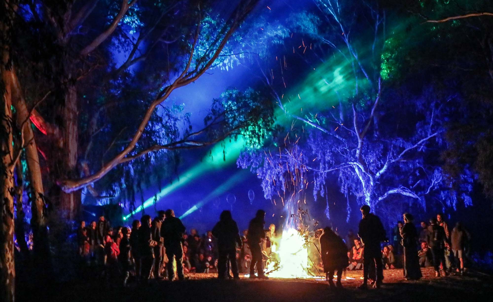 Festival crowd circling a bonfire at night beneath tall trees washed in blue light, green lasers cutting through the canopy