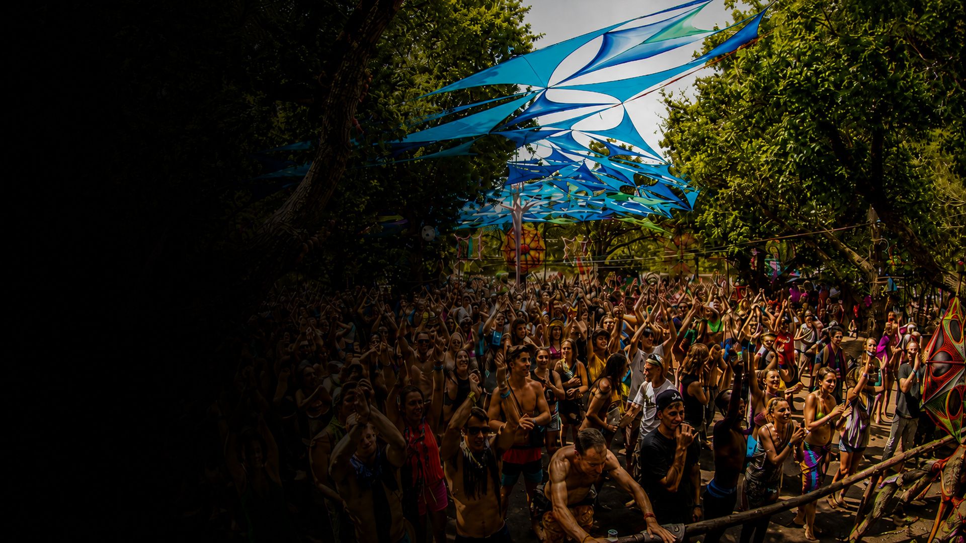A large daylight crowd dancing beneath blue and white shade sails strung between tall trees, with the gathering stretching back into the forest behind them