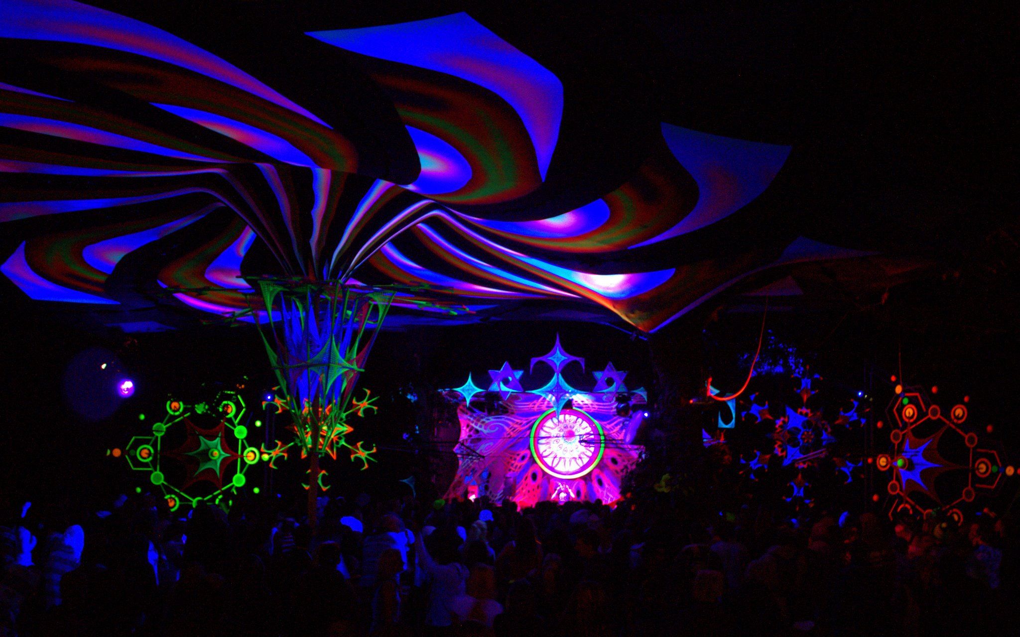Ultraviolet fabric sculptures fanning out above a night crowd facing a lit forest stage
