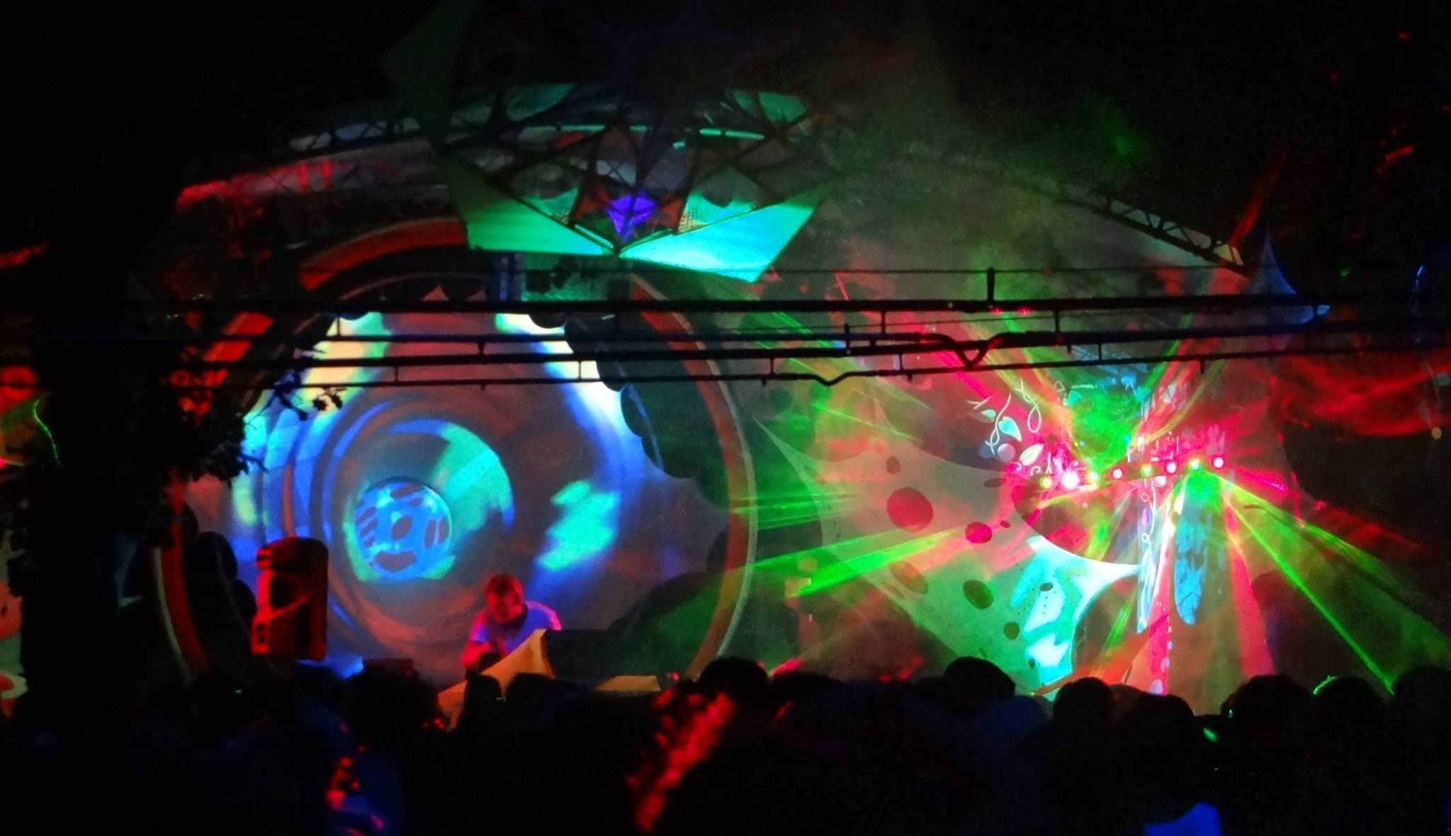DJ playing beneath a projection-mapped stage as green and red lasers fan out over the crowd