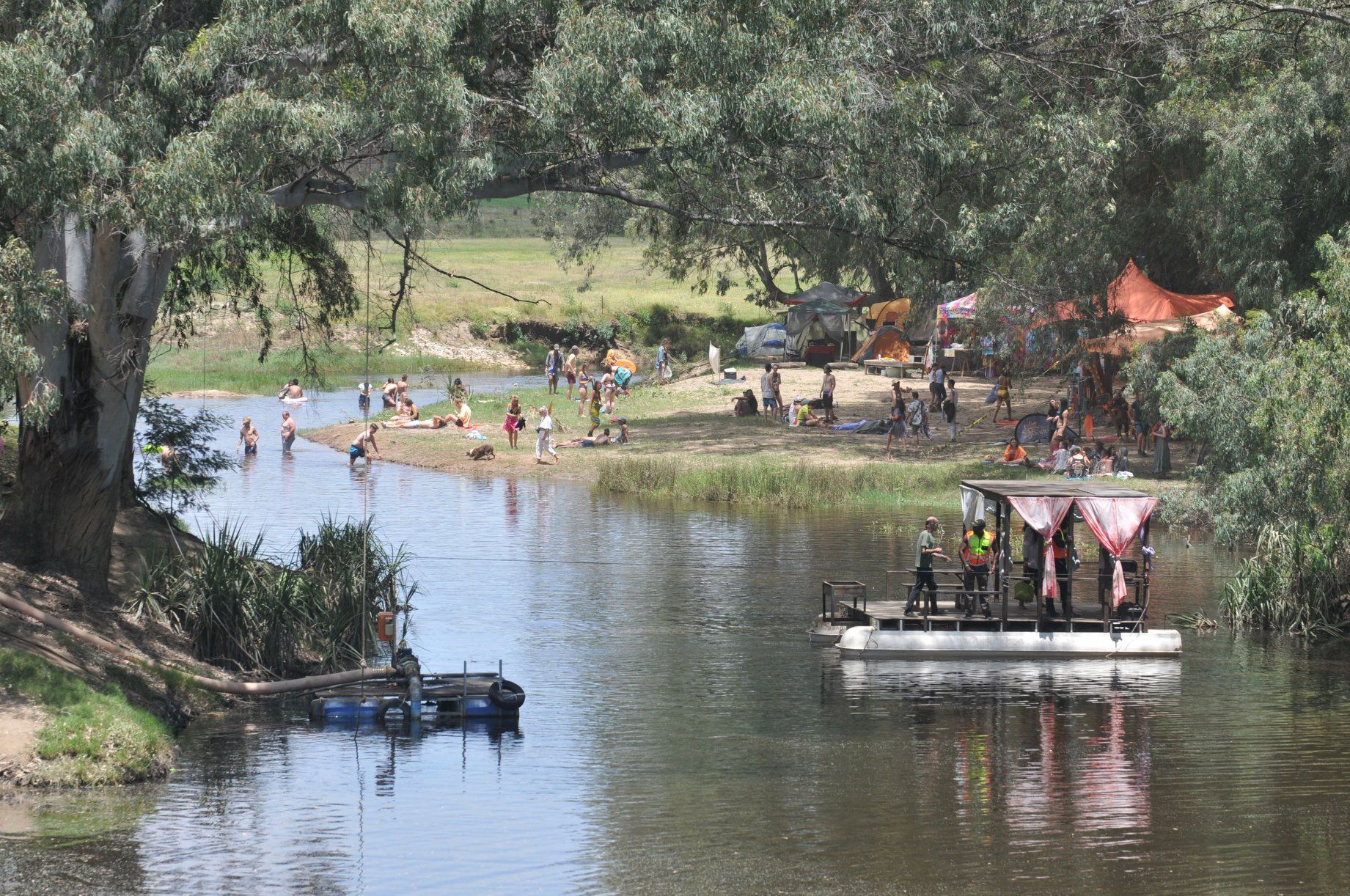 Tents and shade structures pitched among trees along a river bank at a festival camp